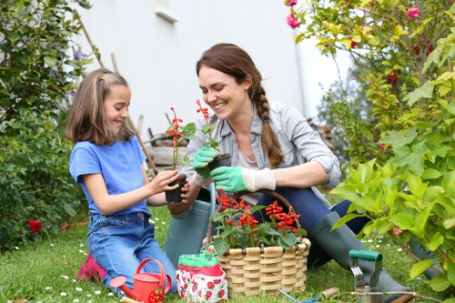 A woman and a young girl are engaged in gardening activities in a lush, well-maintained backyard garden. The woman, dressed casually in a light grey shirt and gardening gloves, is smiling as she holds a small potted plant with red flowers, presenting it to the girl. The girl, wearing a bright blue T-shirt and denim shorts, is seated on the grass and holding the pot with both hands, appearing happy and engaged. The garden features vibrant, leafy green shrubs and plants with pink and red flowering bushes in the background, bordered by a white wall. On the grass nearby, there are gardening tools, a small red watering can, and a woven basket filled with gardening supplies. The scene is set in natural daylight, suggesting a mild, cloudy day suitable for outdoor garden work, and reflects a nurturing outdoor environment typical of a suburban garden in Highgate or similar London area, which emphasizes the importance of gardening services offered by Landscapers Highgate for maintaining such attractive outdoor spaces.