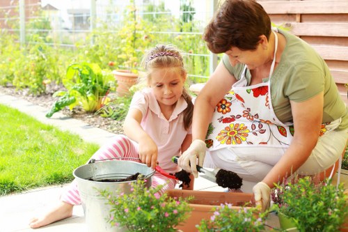 A woman and a young girl are gardening together outdoors in a backyard garden area, with the woman wearing a floral apron and gloves, and the girl in casual clothing with pink and white striped trousers. They are tending to potted plants, with various flowering plants and greenery visible in the foreground and background, including a lush lawn, flower beds, and a wooden fence. The environment shows a well-maintained garden with a paved patio area, and the scene is lit by natural daylight, suggesting a bright, calm day suitable for outdoor gardening activities. This setting demonstrates gardening practices and outdoor maintenance typical of high-quality landscaping services offered by Landscapers Highgate near N6 postcode.