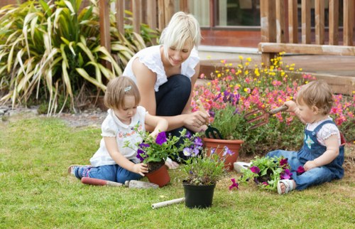 A woman with short blonde hair, wearing a white shirt, is sitting on a well-maintained grassy lawn in a garden, engaged in planting and caring for potted flowers. Two young children, both with light brown hair and wearing casual clothing, are seated on the grass nearby, each tending to their own small pots of colorful flowering plants. The garden features a wooden deck at the back, with a variety of vibrant pink, yellow, and purple flowers planted along its edge, creating a lively and inviting outdoor space. To the left, there are tall, green ornamental grasses adding texture to the garden layout. The overall scene suggests an outdoor gardening activity on a sunny day, with natural light illuminating the scene, emphasizing the lush greenery and bright blooms, aligning with professional gardening and landscaping services offered by companies like Landscapers Highgate in the local Highgate area and the N6 postcode.