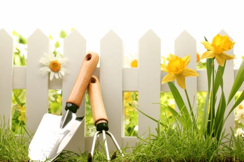 A close-up image of a small, well-maintained garden scene showing a white picket fence in the background, with bright yellow daffodils and white daisies growing along its base. In the foreground, there is a patch of lush green grass with a few garden tools resting on the ground, including a hand trowel with a wooden handle, a pair of garden shears, and a garden fork, suggesting ongoing gardening or maintenance work. The scene is illuminated by natural sunlight, highlighting the vibrant yellow and green colors, indicative of a typical outdoor space in Highgate, suitable for gardening and landscaping enhancement by local professionals like Landscapers Highgate.