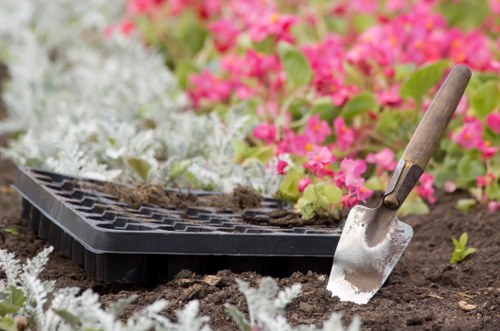 A close-up image of a gardening scene in a landscaped outdoor space, showing a small, silver hand trowel with a wooden handle partially inserted into dark, moist soil. Next to the trowel is a black plastic seed tray filled with soil, resting on the ground. Surrounding the tools are various flowering plants and shrubs, with vibrant pink and white flowers in the background, indicating a well-maintained garden landscape typical of Highgate or nearby areas. The garden features neatly arranged flower beds and soil beds, with a mix of lush green foliage and blooming flowers, suggesting ongoing planting or maintenance work. The lighting suggests a natural outdoor setting, possibly on a mild, overcast day, emphasizing the natural tones and textures of the garden elements, which would benefit from professional gardening or landscaping services from Landscapers Highgate.