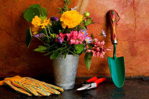 A close-up image of a flowered garden vase placed on a dark stone surface against a textured reddish-brown wall. The bouquet includes large yellow, pink, and purple flowers with green foliage, creating a vibrant display. To the right of the vase, a small green-handled gardening trowel is positioned upright, partially embedded in the surface, with its blade facing downward. Next to the trowel, a pair of red-handled pruning shears lies open, and to the left, a folded pair of brightly striped gardening gloves rests on the surface. The scene is outdoors, suggesting a work station for gardening or landscaping, with natural lighting highlighting the textures of the flowers, tools, and the stone surface, subtly emphasizing skills and care in garden maintenance services provided by landscapers in Highgate or surrounding London areas.