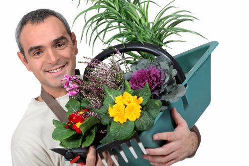 The image shows a smiling man in casual clothing holding a green plastic garden tool caddy filled with a variety of colorful flowering plants such as primroses, violas, and decorative foliage plants, set against a plain white background. Behind him is a lush green potted plant with long, slender leaves, likely a type of house or garden plant, which adds height and structure to the scene. The garden tools and plants are arranged neatly in the caddy, suggesting a focus on gardening tasks such as planting, maintenance, or landscaping. The image emphasizes outdoor gardening activities typical of a well-maintained garden space, which may include lawn care, flower bed tending, and general outdoor plant management. The bright and natural lighting enhances the vivid colors of the flowers and greenery, subtly supporting local garden and landscaping services in areas like Highgate, London, within the N postcode district, and reflecting the professional landscaping work provided by companies like Landscapers Highgate in the local community.