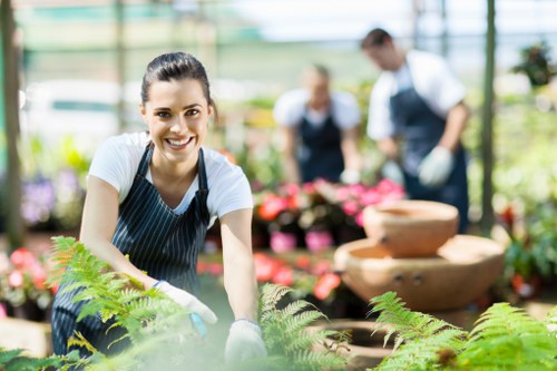 A smiling woman wearing a striped apron and white gloves is in the foreground of a greenhouse, tending to lush green ferns and other plants in a garden center or nursery. Behind her, two blurred individuals are working with flowering plants positioned on tiered wooden display tables. The greenhouse features large glass panels allowing natural light to illuminate the vibrant plants, with a variety of flower pots and trays visible. The scene showcases a well-maintained outdoor workspace focused on gardening and plant care, with a backdrop of additional greenery and flowering plants, suggesting an environment that supports landscape and garden outdoor maintenance services provided by companies like landscapershighgate.co.uk in the Highgate area.